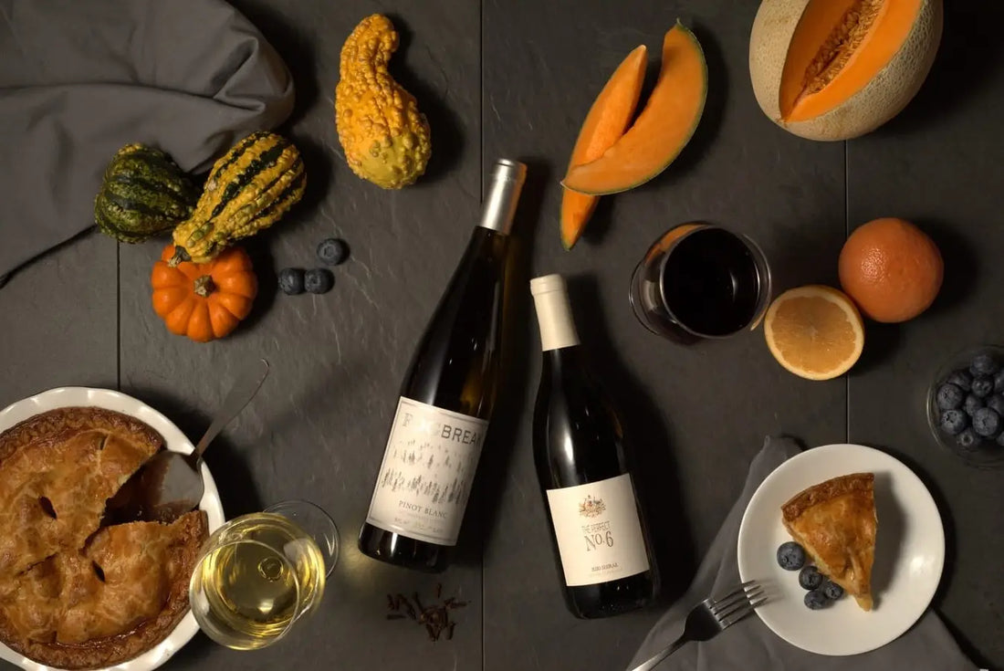 Two bottles of red wine, apple pie, gourds, melon, and oranges.