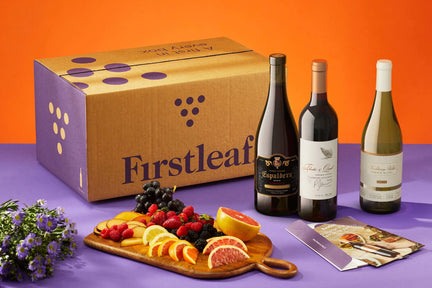 Three bottles of Firstleaf wine on a purple table with an orange background; a Firstleaf box and a fruit platter.