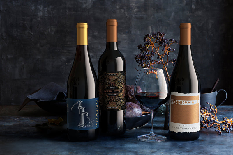 Three bottles of Firstleaf Fine Wine on a dark background with a glass of red wine, flowers, and plums.