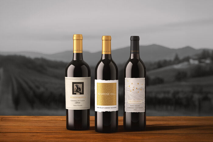 Three wine bottles on a wooden surface with a blurred vineyard background