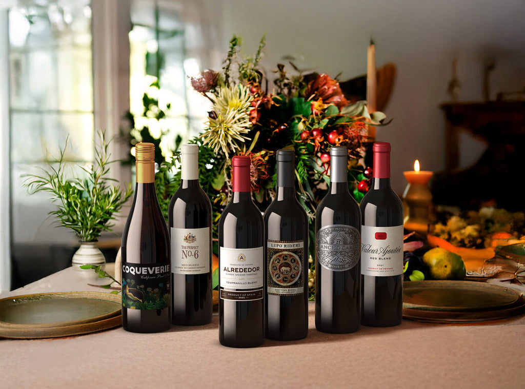 Six bottles of red wine on a table with flowers and candles.