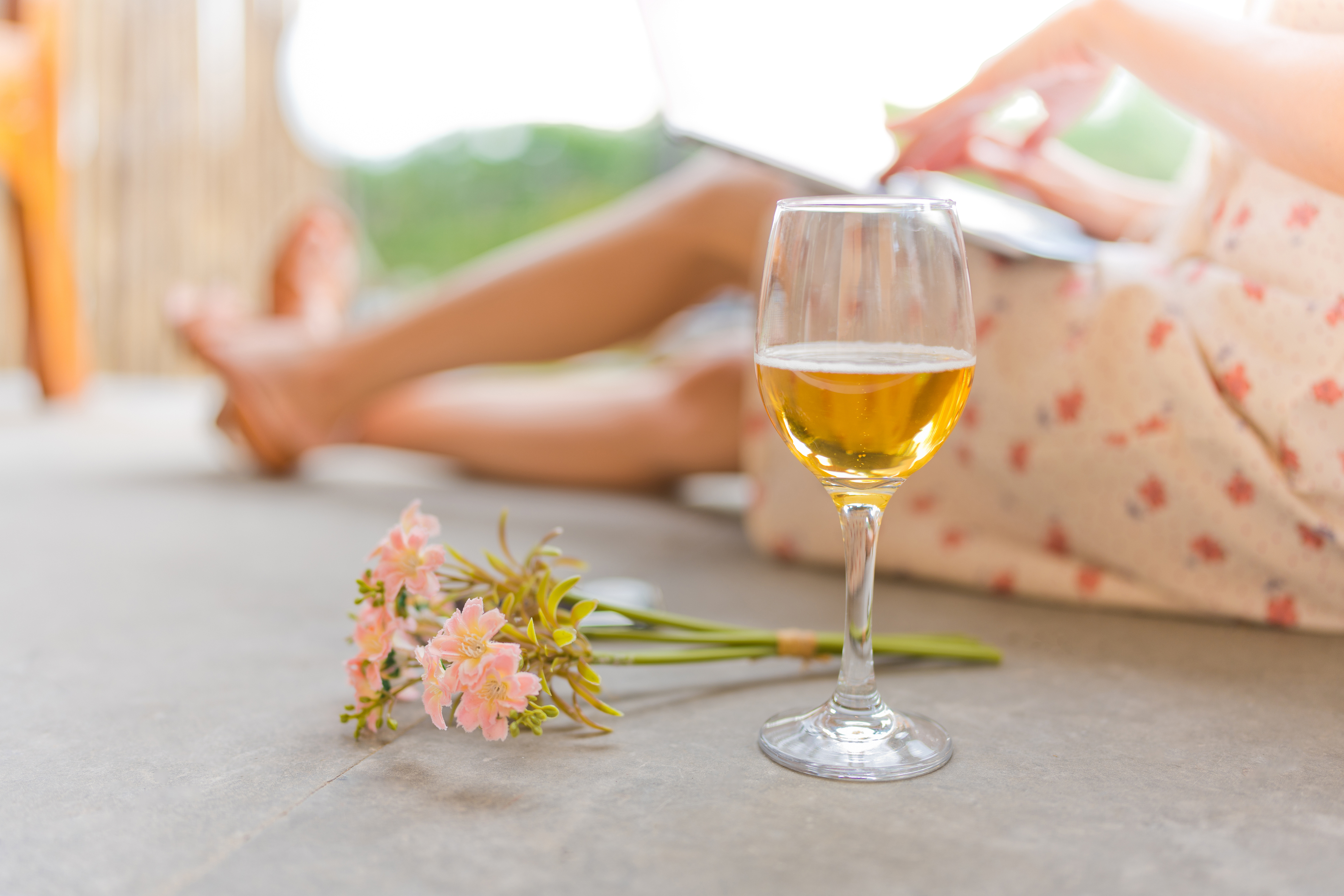 Image of a glass of white wine and pink flowers with a woman reading a book in the background