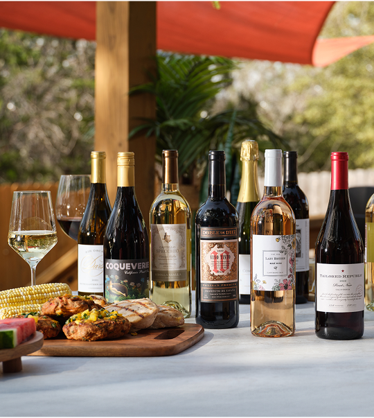 Image, Several bottles of Firstleaf wine on an outdoor table next to meat, corn, and fruit.