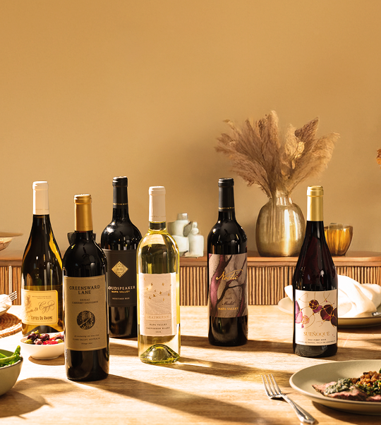 A wooden dining table set with six bottles of wine, including both red and white varieties, surrounded by plates, glasses, and simple table décor in warm lighting.