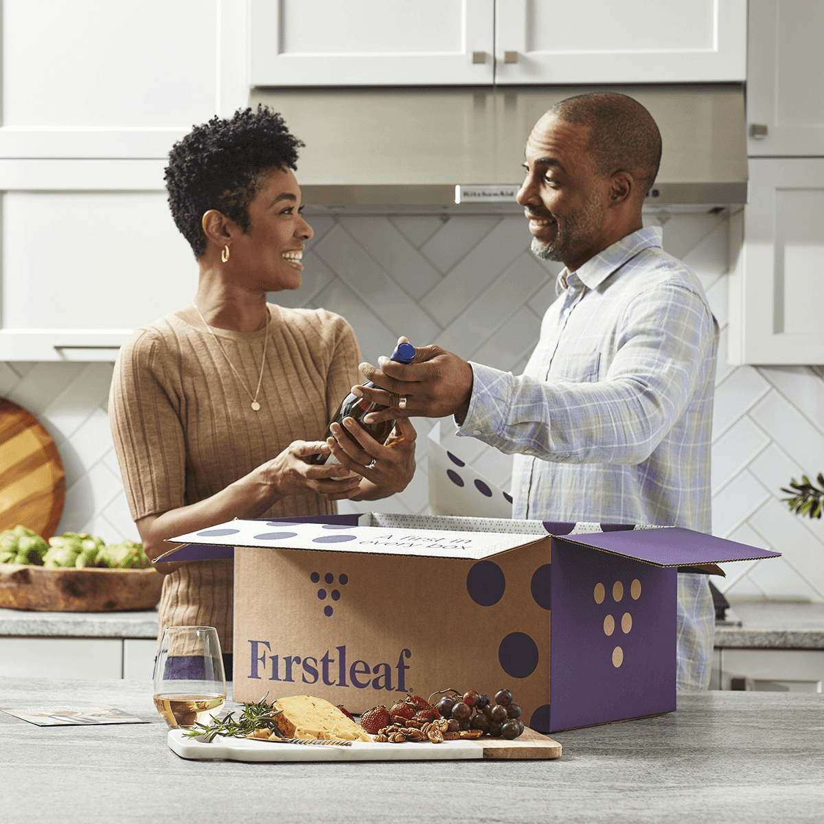 Smiling couple unboxing wine bottles from a Firstleaf delivery box in a modern kitchen, with a wine glass and charcuterie board on the counter.
